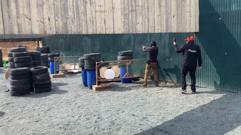 Salvador Sanpascual tira con pistola junto a una pared de chapa verde, entre torres de neumáticos y bidones azules, con otra persona detrás.